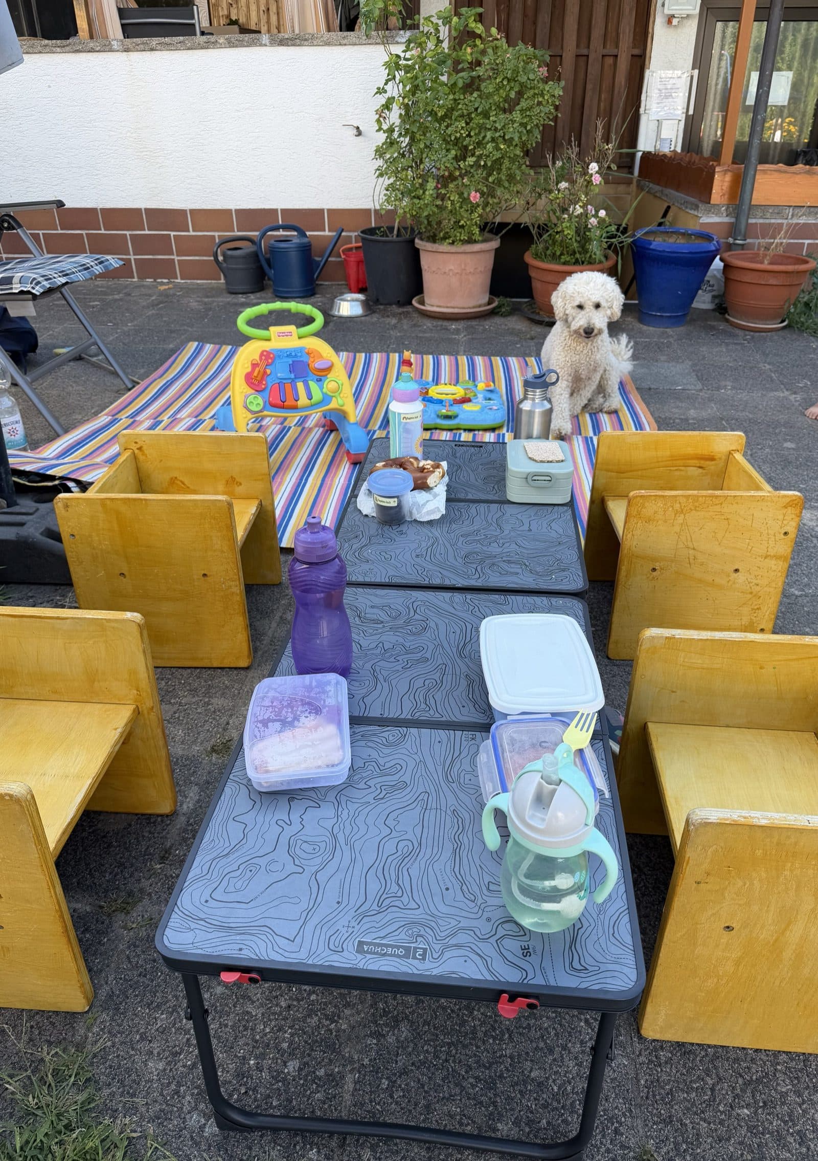 Der gedeckte Kindertisch im Garten mit kleinen Holzstühlen, im Hintergrund Pudeldame Lucy