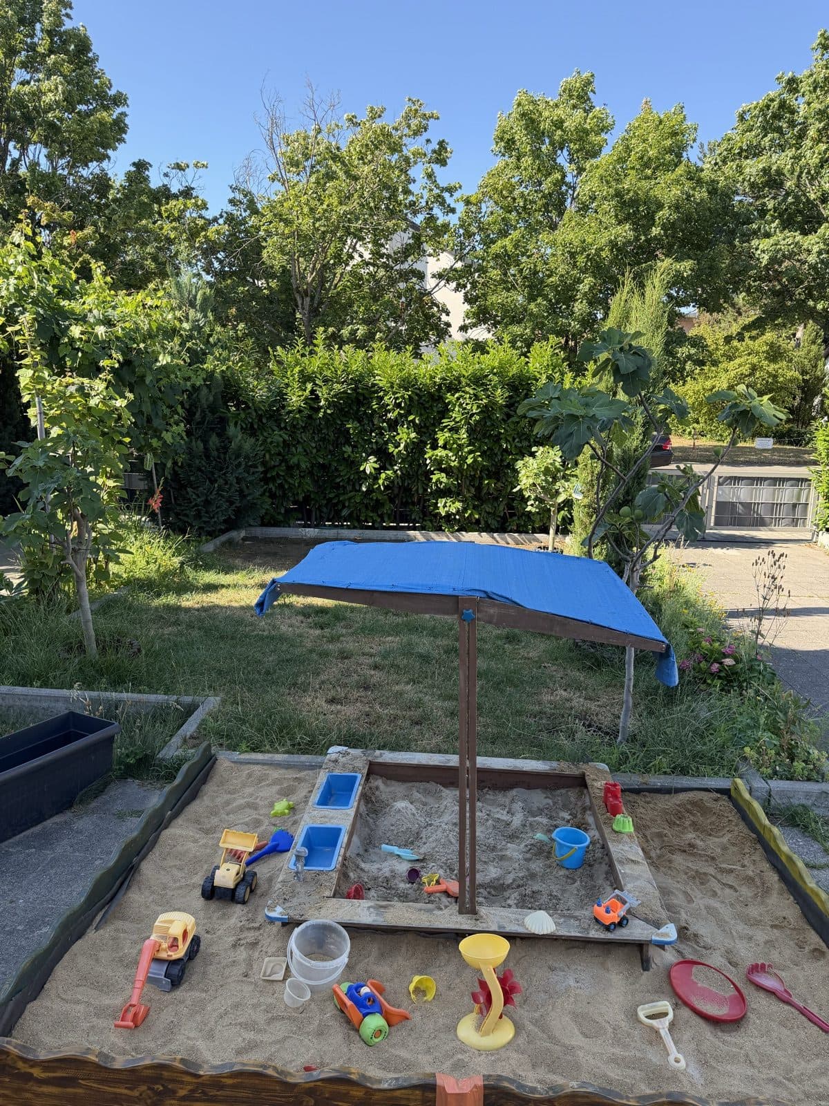 Der Garten von Tagesmutter Nilo mit überdachtem Sandkasten, Wiese und hohen Bäumen